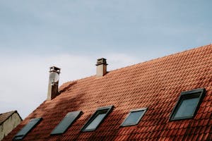 A picturesque red tiled roof featuring chimneys and skylight windows against a blue sky.