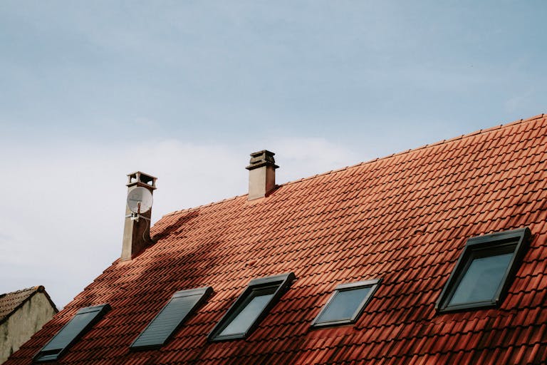 A picturesque red tiled roof featuring chimneys and skylight windows against a blue sky.